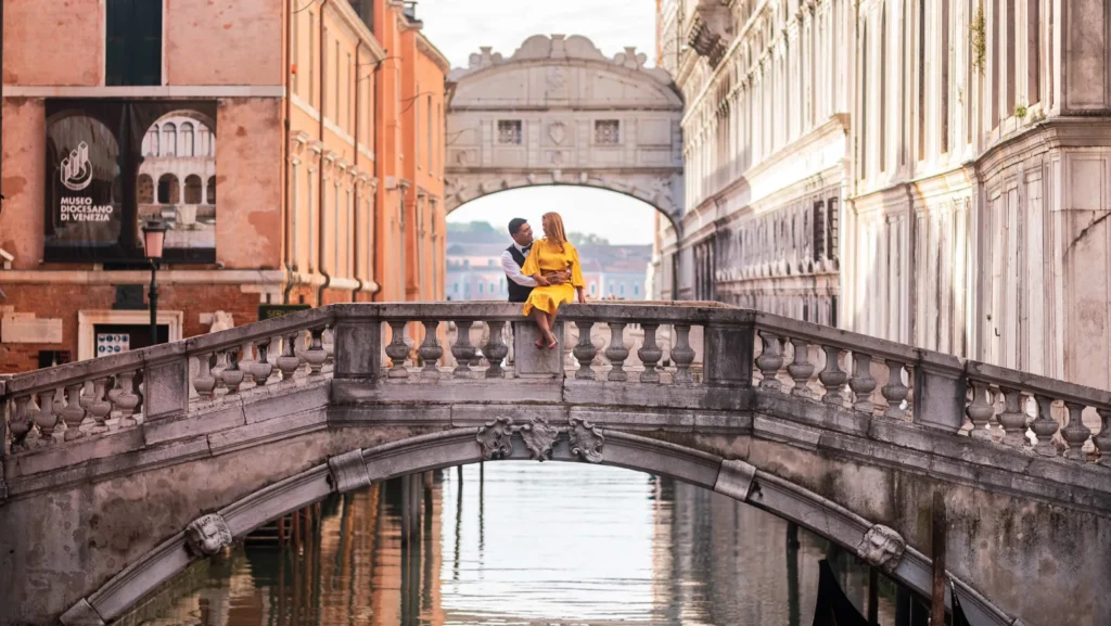 Engagement Venice photo of a couple sitting on a stone bridge near Doge’s Palace with the Bridge of Sighs in the background at sunrise